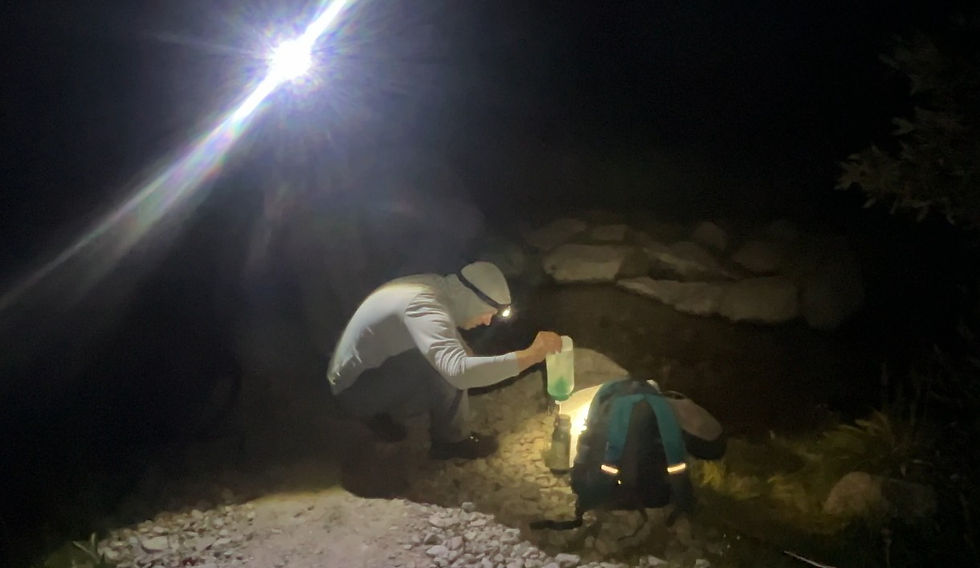 Man collecting water at night by a stream with a headlamp. Backpack and lantern nearby. Dark, rocky setting with focused light.