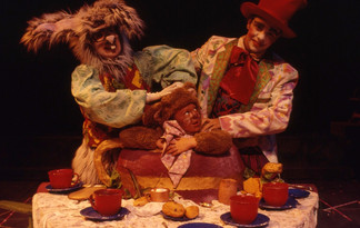 In a vibrant tea party scene, the March Hare (Kathy Ferman) and the Mad Hatter (Jeremy Williams) laugh over the Dormouse (Melissa Riggins), who is tucked inside a large teapot wearing fuzzy brown pajamas and a napkin bib. The table in front of them is cluttered with teacups, saucers, leaves, and pastries. Their painted faces and expressive costumes highlight the surreal, joyful madness of the scene.