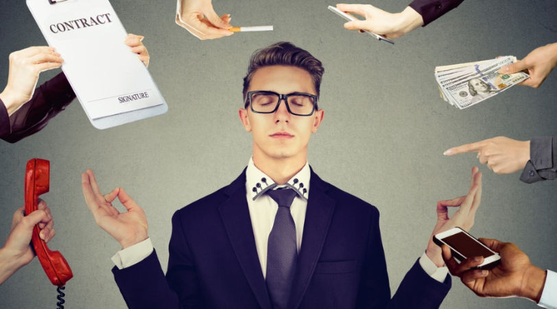 Young adult man in a suit meditates, surrounded by hands offering a contract, phone, money, cigarette. The man displays a calm mood, despite 6 hands around him offering something that requires his attention. The image has a gray background, business theme.