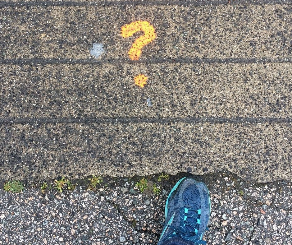 Image from perspective from above, looking down at a sidewalk. Blue sneaker on a textured sidewalk with orange question mark painted next to a silver spot on the sidewalk. Sparse greenery in sidewalk cracks, conveying curiosity.