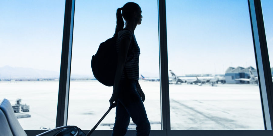 Silhouetted young woman with a backpack and suitcase walks in an airport terminal, large windows reveal planes on the tarmac, blue sky. The woman is looking out the windows to the planes on the tarmac.
