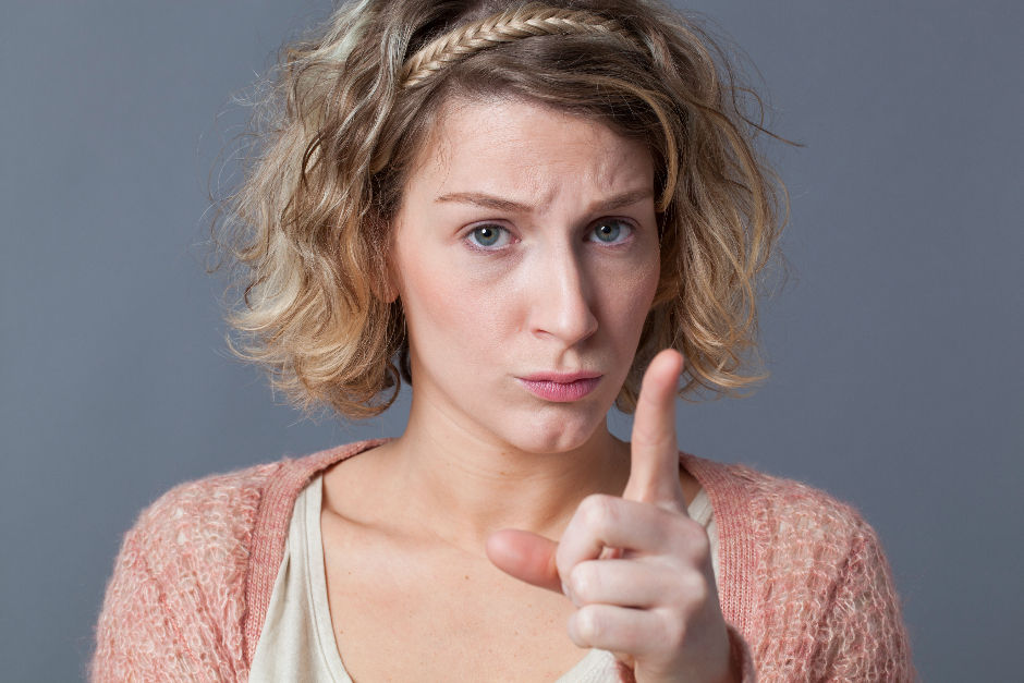 Woman with short, curly, light brown hair and a braided headband points sternly. She wears a pink cardigan against a gray background, conveying seriousness and providing scolding or telling the view what they should be doing.