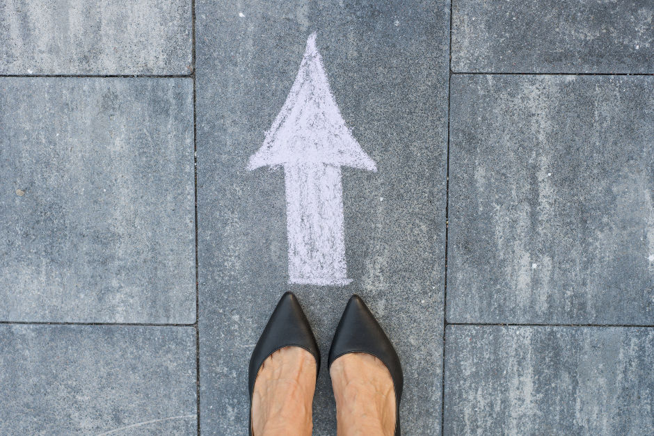 Perspective viewpoint of looking down at feet wearing black pointed women's shoes on gray pavement with a white chalk arrow pointing forward, suggesting direction or progress.