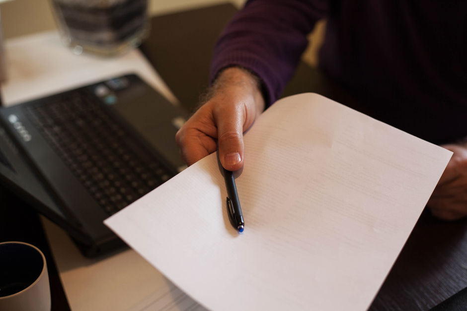 Hand offering a pen and paper over a laptop on a desk, as if requesting to have a contract or agreement signed. Person wears a purple sweater. Warm lighting, suggesting a serious or formal setting.