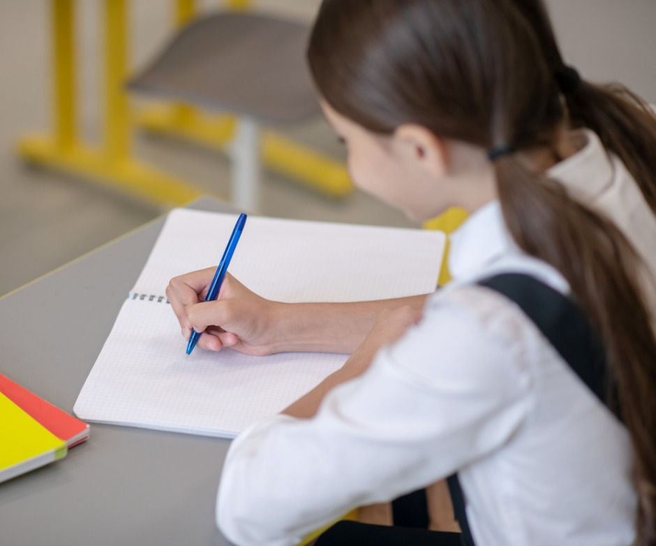 Girl in white shirt writing with a blue pen on a notebook at a desk in a school. Yellow and red folders are nearby. Classroom setting, focused mood.