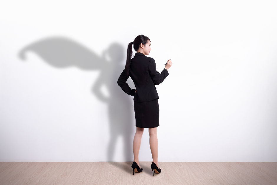 A woman in a black business suit stands confidently. Her shadow on a white wall resembles a superhero with a cape. Bright, minimalist setting.