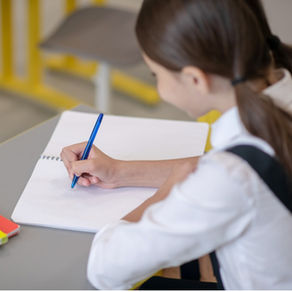Girl in white shirt writing with a blue pen on a notebook at a desk in a school. Yellow and red folders are nearby. Classroom setting, focused mood.