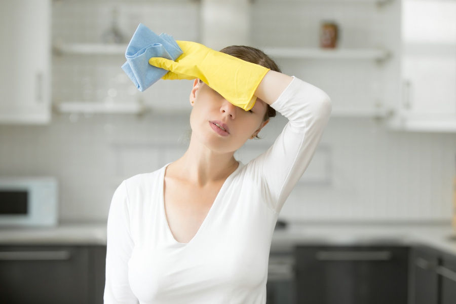 Woman in a kitchen with yellow gloves and a blue cloth, wiping her forehead. She appears to be doing housework and looks tired. Background has white cabinets and shelves.