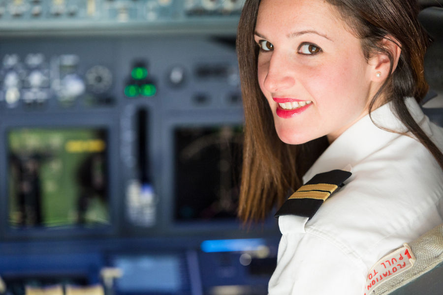 Smiling female pilot in white uniform with epaulettes seated in airplane cockpit, colorful control panel in background, relaxed mood.
