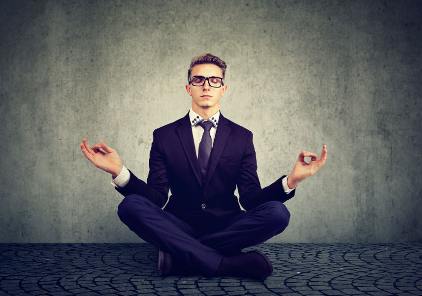 Young adult man in a suit sitting cross-legged, meditating with closed eyes. Concrete background, cobblestone floor. Calm and focused mood.