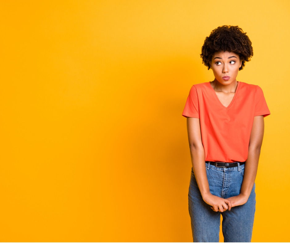 Woman in orange shirt and jeans looks curious with a hesitant and unsure expression against a bright yellow background.