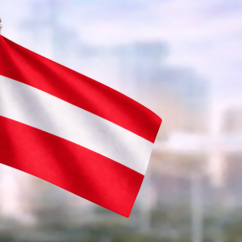 Austrian flag in red and white stripes, photographed in close-up against a softly blurred urban landscape.