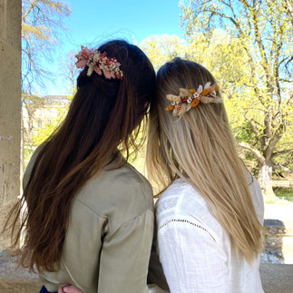 Photographie de deux personnes de dos portant un peigne floral dans leur coiffure. La personne de gauche porte un haut kaki et a les cheveux bruns. Celle de droite un chemisier blanc et a les cheveux blonds. Cette photographie a été prise pour mettre en valeur l’accessoire floral dans leur coiffure.