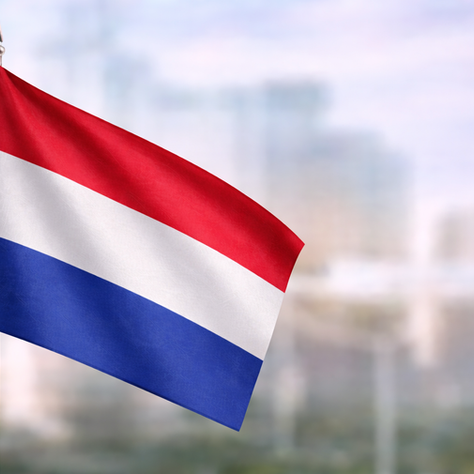 Dutch flag in red, white and blue waving in close-up, set against a softly blurred urban backdrop in daylight.