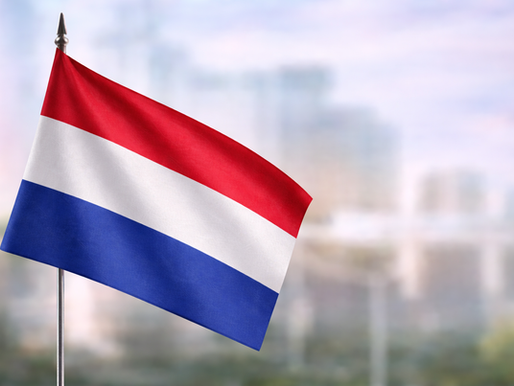 Dutch flag in red, white and blue waving in close-up, set against a softly blurred urban backdrop in daylight.