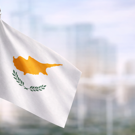 Cyprus flag with white background, golden island silhouette and olive branches, shown in close-up with a blurred city background.