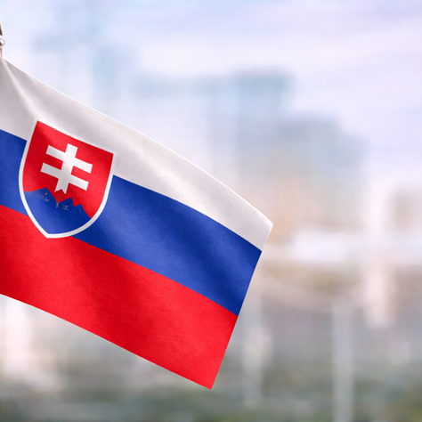 Slovak flag featuring white, blue and red stripes with national emblem, shown in close-up against a soft city background.e 