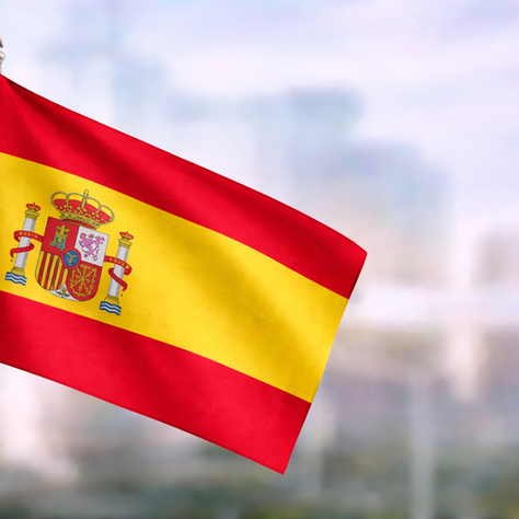 Spanish flag with red and yellow stripes and coat of arms, shown in close-up with a soft-focus city background.