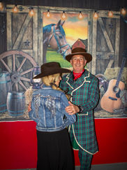 A couple in western attire smiles while dancing; the man wears a plaid coat and red scarf