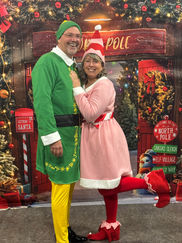 A man and woman dressed in playful elf costumes posing together in front of a North Pole sign