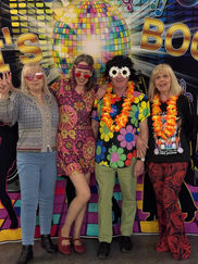 Four people in vibrant 70s attire stand joyfully on a colorful disco floor. They wear flower patterns