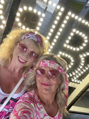Two women wearing pink, retro-themed outfits and matching sunglasses pose happily in front of a lit sign
