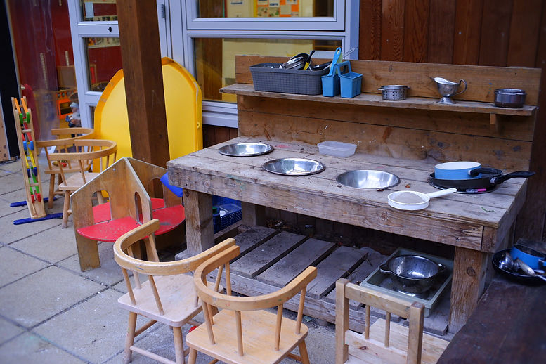 The outdoor play kitchen, which has a large wooden structure with metal bowls sunk in, pans, colanders and other equipment, some child-height wooden chairs. The garden is accessed from a door on the right-hand side of the main hall.
