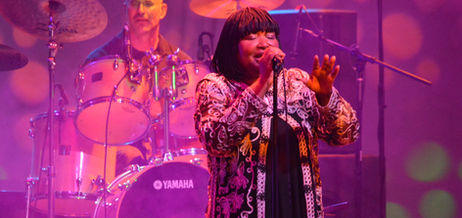 Hazel Miller singing with her arm outstretched with a misty pink background.