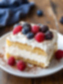 A slice of Tres Leches Cake on a white plate, drizzled with sweetened condensed milk. The cake is moist and fluffy, with a golden brown top. Whipped cream and fresh berries decorate the top, and a dusting of cinnamon adds a touch of warmth. The background is a rustic wooden table, with a shallow depth of field to focus on the cake.