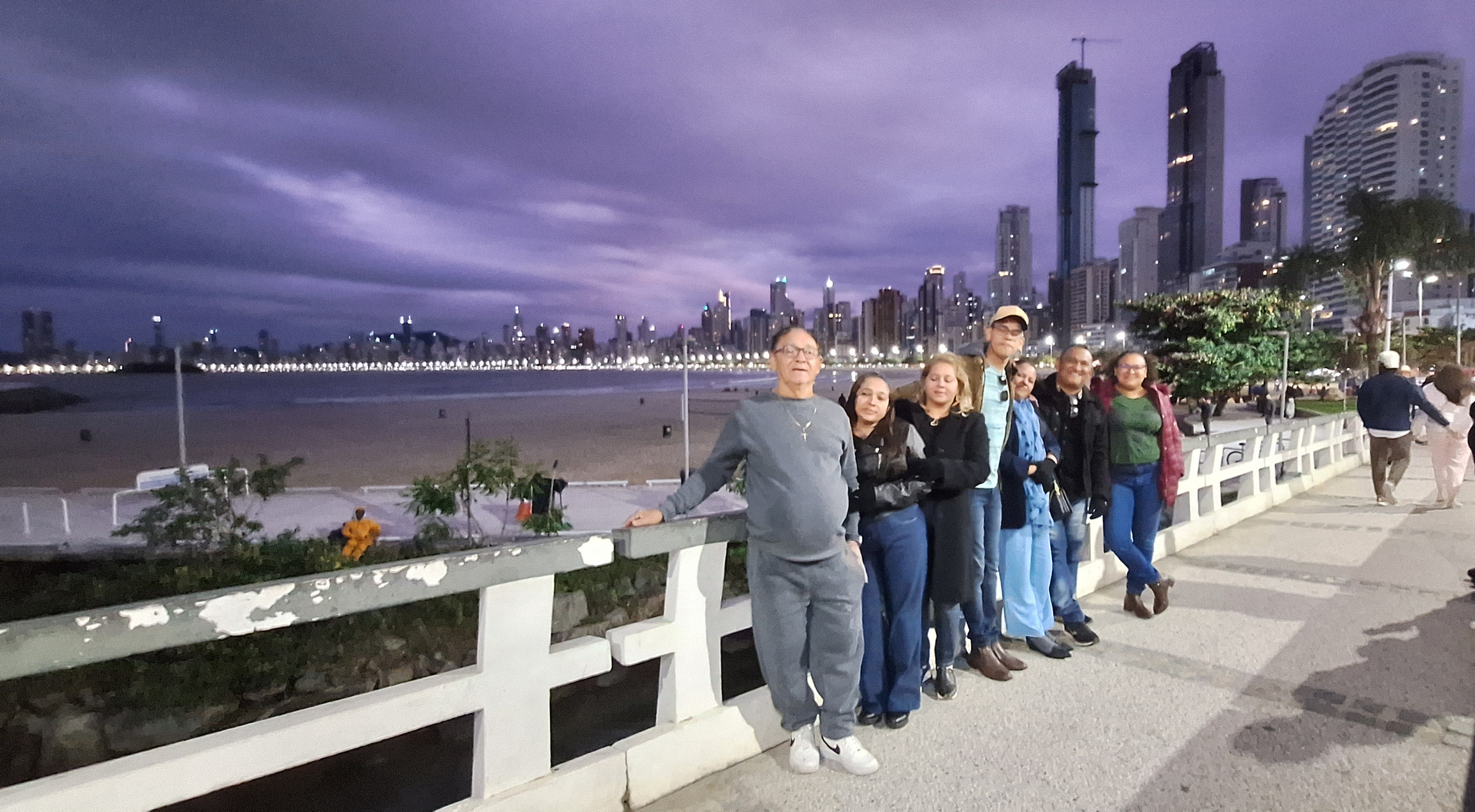 Grupo de turistas na orla de Balneário Camboriú ao entardecer, com vista dos arranha-céus da Dubai brasileira