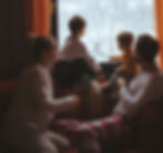 Family in pajamas sits by a window, watching snow outside. Warm colors inside, snowy landscape visible. Cozy, relaxed atmosphere.
