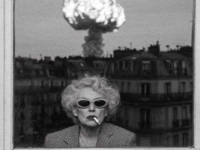 Mulher de óculos escuros e cigarro na janela, com cogumelo nuclear ao fundo sobre a cidade em preto e branco.