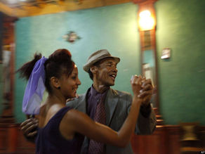 Duas pessoas dançam sorrindo em um salão de cor verde. O homem usa chapéu e terno; a mulher, vestido roxo com fita no cabelo.