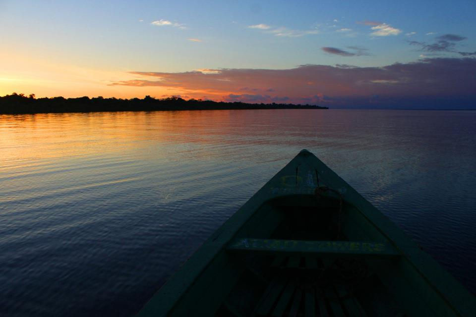 A green canoe moves along the river, reflecting the orange sunset. The sky transitions from orange to dark blue, creating a serene atmosphere.