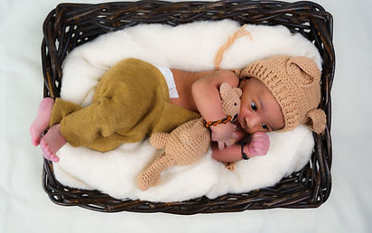 newborn baby curled up in a blanket filled basket wearing knitted hate and dungarees 