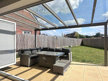 Silver bronze frame aluminium veranda with a glass roof, furnished with beige rattan garden sofas and a glass dining table, designed as a lounge and dining area.