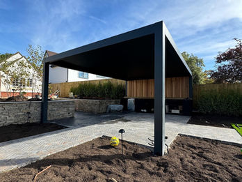 Anthracite grey aluminium frame louvered pergola with a timber wall, kitchen cabinets, grill and sink. Furnished with grey steel and wooden outdoor dining chairs and table.