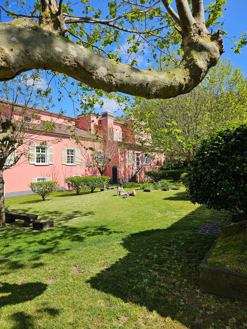 Shaded garden area at Solar de Lalem with lush grass, stone benches, and the estate’s signature pink building partially framed by a large tree branch under a bright blue sky