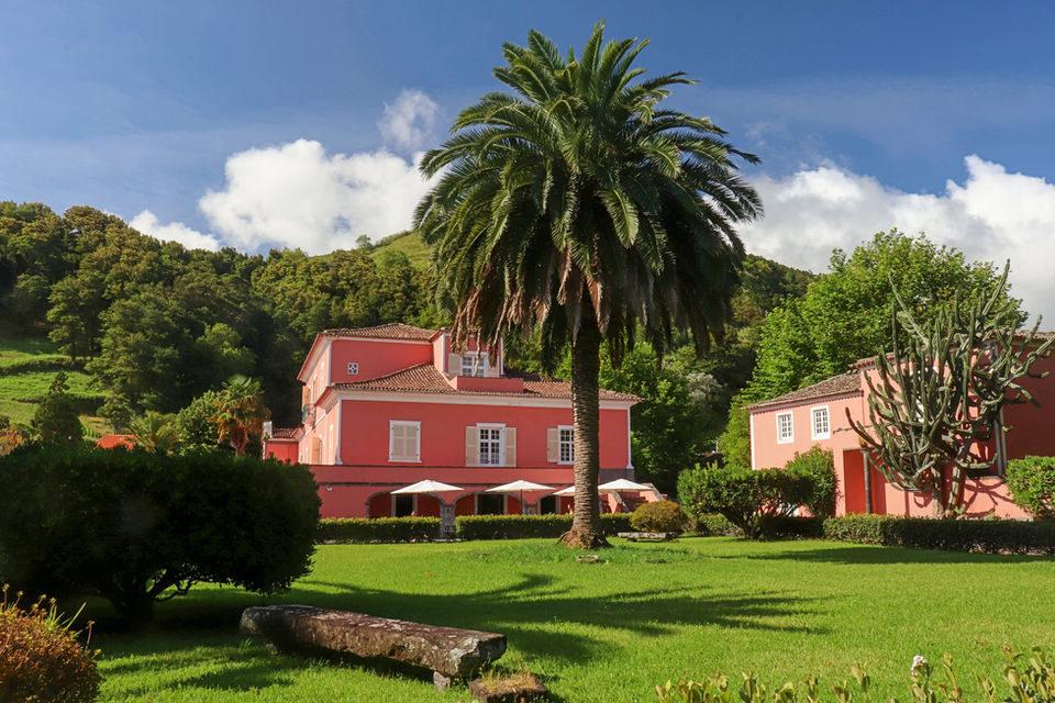 Front view of Solar de Lalem with its striking pink facade, centered around a tall palm tree and surrounded by manicured lawns, lush greenery, and forested hills under a sunny sky