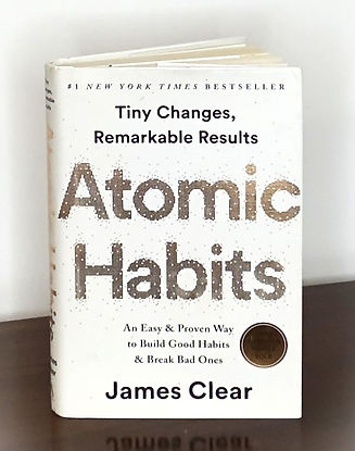 Book sitting on a brown surface with grey background. Book title is Atomic Habits by James Clear.