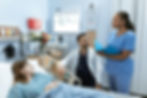 A hospital room with female patient in a bed, a male healthcare provider seated by the bedside and a female healthcare worker holding a folder standing by the bed. There is an eye chart hanging on the wall along with an x-ray image reader, sharps container, blood pressure cuff.