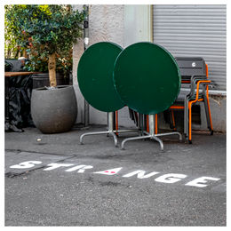 Tables and chairs in front of a closed restaurant and the word STRANGE written on white on the pavement