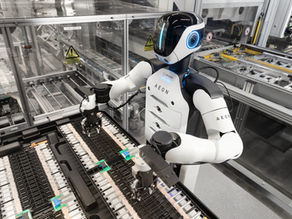 Humanoid AEON robot with glowing blue face works on a factory conveyor, handling parts inside a glass-enclosed assembly line.
