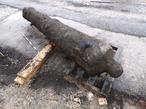A weathered cannon rests on a wooden pallet on a rough concrete surface. A measuring rod is beside it, indicating it's part of an excavation.
