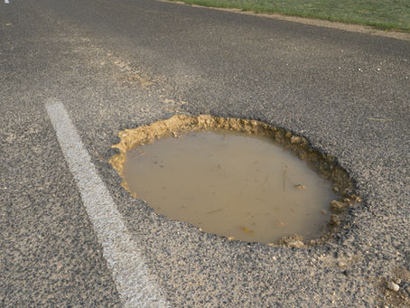 Large pothole filled with muddy water on an asphalt road beside a white lane line and grassy shoulder.