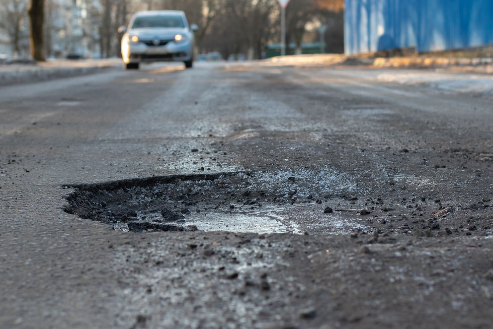 Close-up of a deep pothole on an asphalt road with a car approaching. Background shows a blurred blue wall and bare trees.