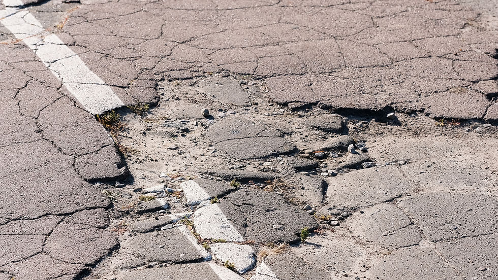 Cracked, crumbling asphalt road with faded white lane markings and potholes under bright sunlight.