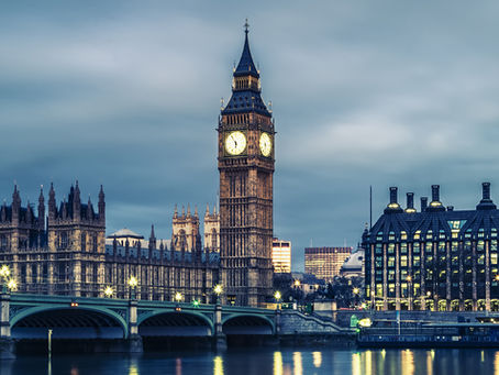 Clock tower and lit buildings by a river at dusk, under a cloudy sky. A bridge crosses the calm water, reflecting lights. Moody ambiance.