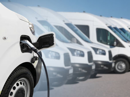White electric van charging in the foreground, with a line of similar vans parked in the background under a clear blue sky.