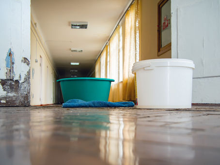 Buckets and cloth on wet floor in a dim hallway with peeling paint and yellow curtains, creating a neglected, sombre mood.
